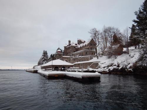 A snow-covered stone mansion by the water, surrounded by trees, with a dock extending into the calm lake.