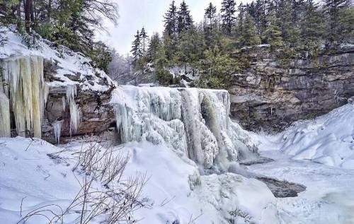A frozen waterfall surrounded by snow-covered rocks and evergreen trees in a winter landscape.