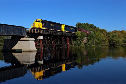 A yellow and blue train crosses a bridge over a calm river, reflecting the scene in the water below.