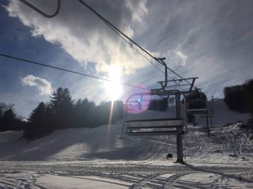 A ski lift against a snowy landscape, with the sun shining through clouds in the background.