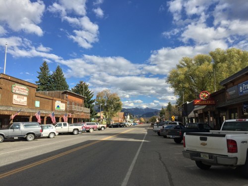 A small town street lined with shops, parked trucks, and mountains under a blue sky with fluffy clouds.