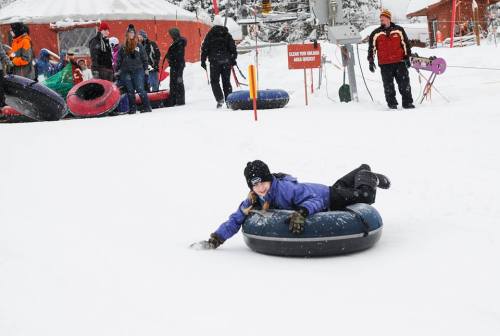 A girl in a purple jacket lies on a snow tube, enjoying a snowy tubing hill with others in the background.