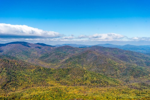 A panoramic view of rolling mountains covered in autumn foliage under a clear blue sky with scattered clouds.
