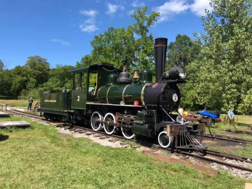 A vintage steam locomotive on tracks, surrounded by greenery and blue skies, with a person working nearby.