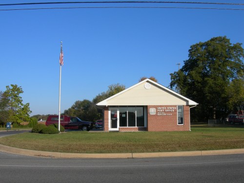 A small brick post office building with a flagpole, surrounded by trees and a clear blue sky.