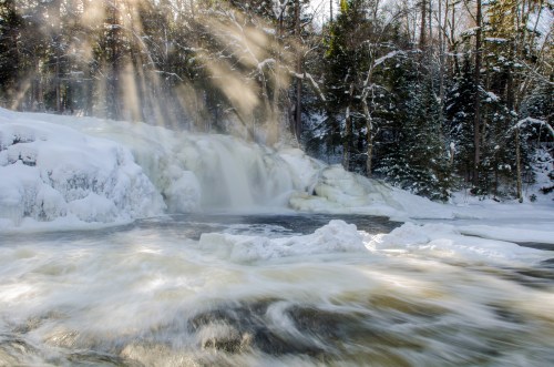 A snowy waterfall cascades into a river, with sunlight streaming through trees, creating a serene winter scene.