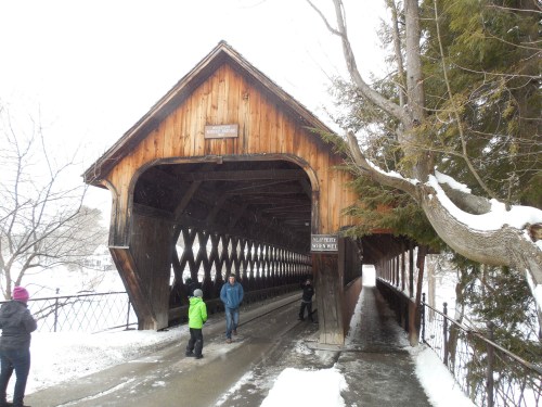 A snowy scene featuring a wooden covered bridge with people walking through it, surrounded by trees.