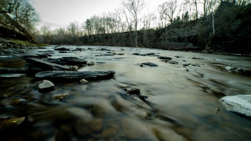A serene river flows over smooth stones, surrounded by trees and a calm, twilight sky.