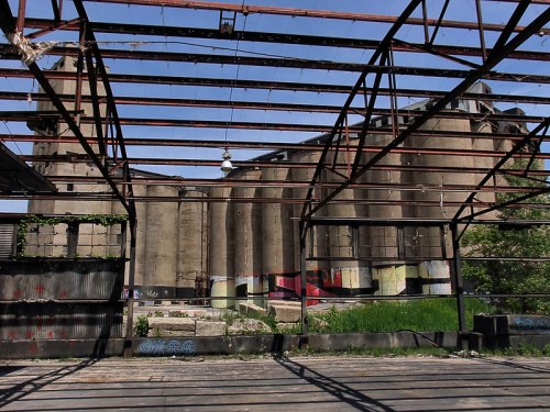 Abandoned industrial building viewed through a rusted metal frame, with greenery and graffiti visible.