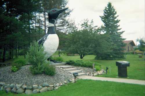 A large statue of a loon stands in a grassy area, surrounded by trees and shrubs, with a path leading up to it.