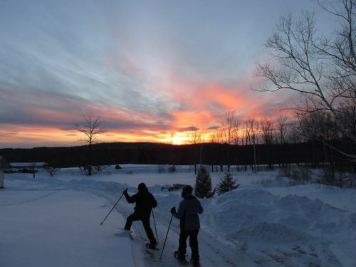Two people snowshoeing at sunset, with a colorful sky and snow-covered landscape in the background.