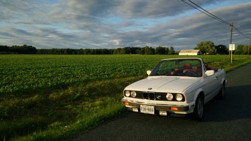 A white convertible BMW parked on a rural road beside a green field under a cloudy sky.