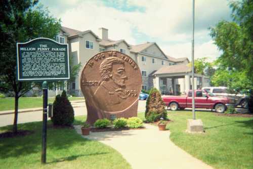 A large penny sculpture stands in front of a historical sign and residential buildings, with a blue sky above.