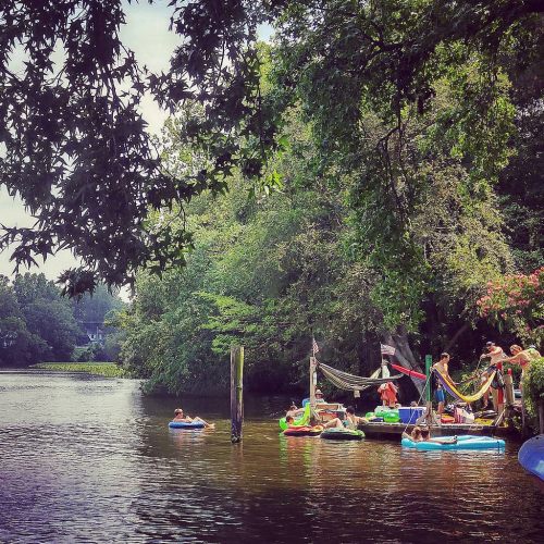 A serene river scene with people floating on tubes and relaxing on hammocks among lush greenery.