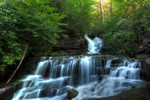 A serene waterfall cascades over rocks, surrounded by lush green trees and dappled sunlight filtering through the leaves.