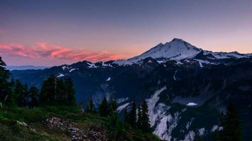 A stunning mountain landscape at sunset, featuring snow-capped peaks and colorful clouds above a lush forest.