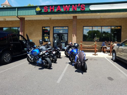 A row of motorcycles parked outside Shawn's restaurant, with diners visible on the patio. Sunny day.