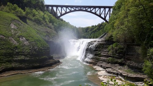 A scenic view of a waterfall cascading over rocky cliffs, with a bridge arching above and lush greenery surrounding the area.