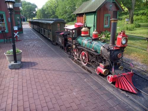 A vintage steam locomotive with a red and green color scheme, parked at a quaint train station surrounded by greenery.