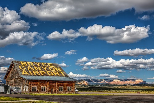 A rustic wooden building with "Bryce Canyon Airport" sign, set against a backdrop of mountains and a blue sky with clouds.