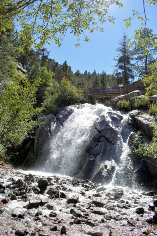 A waterfall cascades over rocks, surrounded by lush greenery and trees under a clear blue sky.