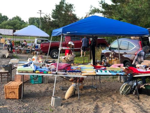 A bustling outdoor market with tables of goods under blue tents, surrounded by vehicles and shoppers.
