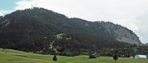 A lush green hillside with a mountain in the background, featuring trees and a small building in the foreground.
