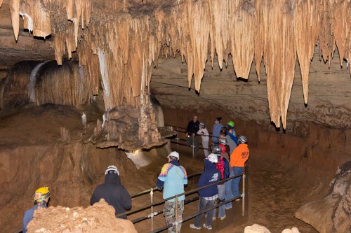 A group of people wearing helmets explores a cave with stalactites and a small water feature.