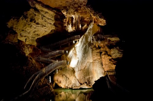 A dimly lit cave with illuminated rock formations and winding stairs leading down to a reflective pool of water.