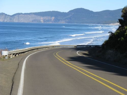 A winding road curves along a coastline with waves crashing on the shore and mountains in the background.