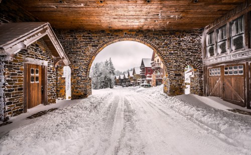 Snow-covered street framed by a stone archway, with rustic buildings and trees in a winter landscape.