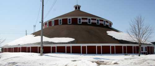 A large, round building with a brown roof and red walls, surrounded by snow on a clear day.