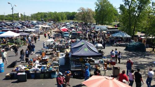A bustling outdoor flea market with numerous stalls, tents, and crowds of people browsing various items under a clear blue sky.