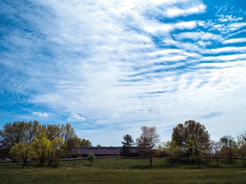 A clear blue sky with wispy clouds above a green field and trees, with a building in the background.