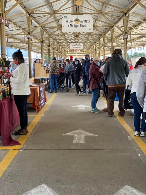 A bustling market scene with people browsing stalls under a covered walkway, following directional arrows on the ground.