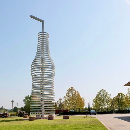 A large, spiraled sculpture resembling a bottle with a straw, set in a grassy area under a clear blue sky.