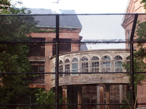 Abandoned brick building with arched windows, partially obscured by a black fence and surrounded by greenery.