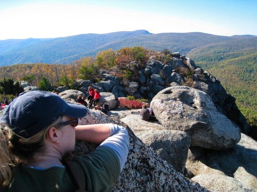 A person leans on a large rock, overlooking a scenic mountain view with colorful autumn foliage and hikers in the background.