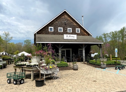 A charming garden center with a wooden building, surrounded by flower displays and decorative carts on a paved path.
