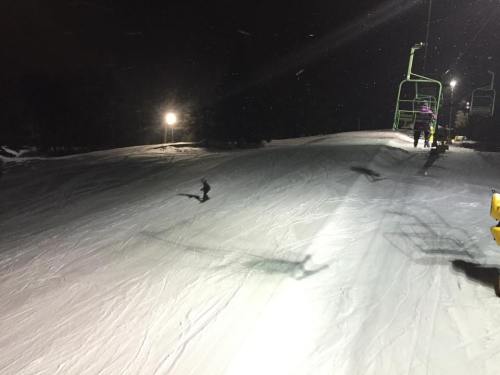 A snowy ski slope at night, with a ski lift and a few people in the background under bright lights.
