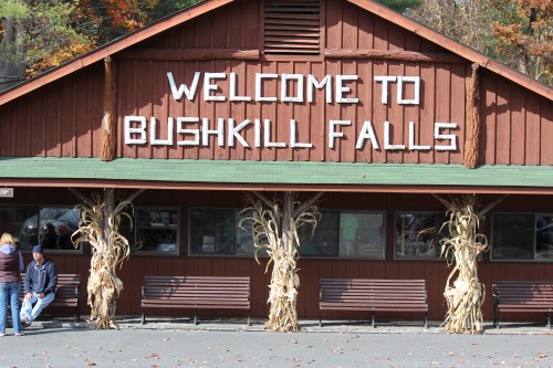 A wooden building with a sign reading "Welcome to Bushkill Falls," decorated with corn stalks on either side.