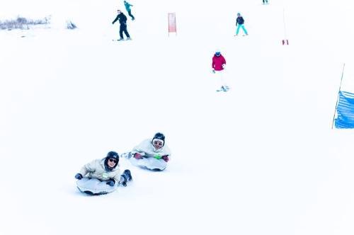 Two people sit on sleds in the snow, while others ski and play in the background on a snowy hill.