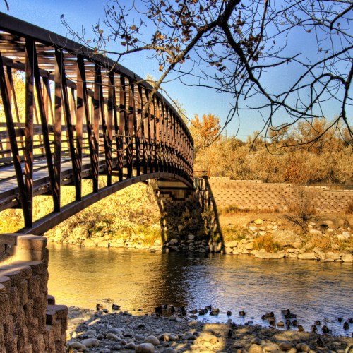 A curved wooden bridge spans a calm river, surrounded by trees and rocky banks under a clear blue sky.