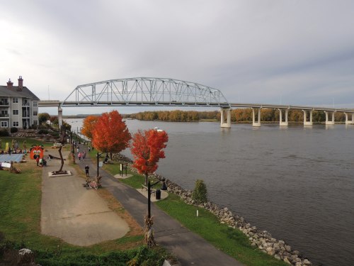 A river scene featuring a bridge, autumn trees, and a pathway with sculptures along the water's edge.