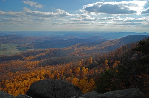 A panoramic view of rolling hills covered in autumn foliage, with clouds and sunlight illuminating the landscape.
