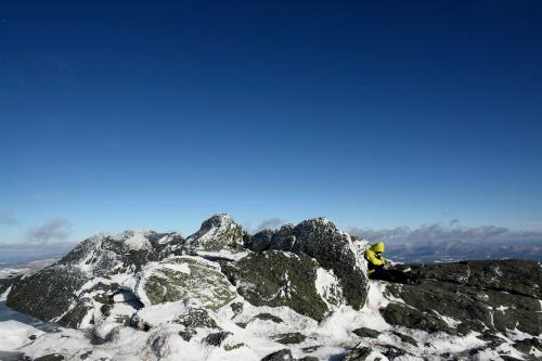 A person in a yellow jacket sits on rocky, snow-covered terrain under a clear blue sky.