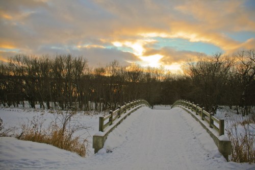 A snow-covered bridge leads into a winter landscape, with trees silhouetted against a colorful sunset sky.