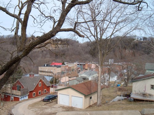 A view of a small town nestled in a valley, with houses, trees, and distant hills under a cloudy sky.