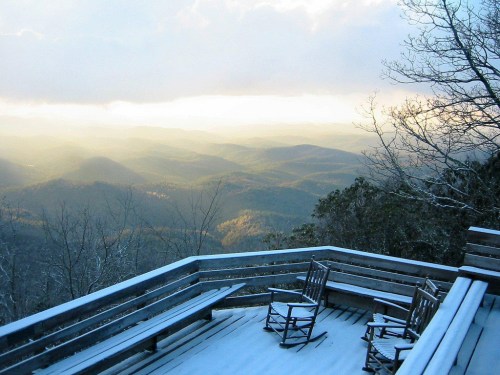 Snow-covered deck with two rocking chairs overlooking misty mountains and a cloudy sky.