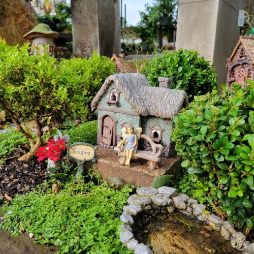 A miniature garden scene featuring a small house, a bench with a figure, and lush greenery. A sign reads "Imagine."